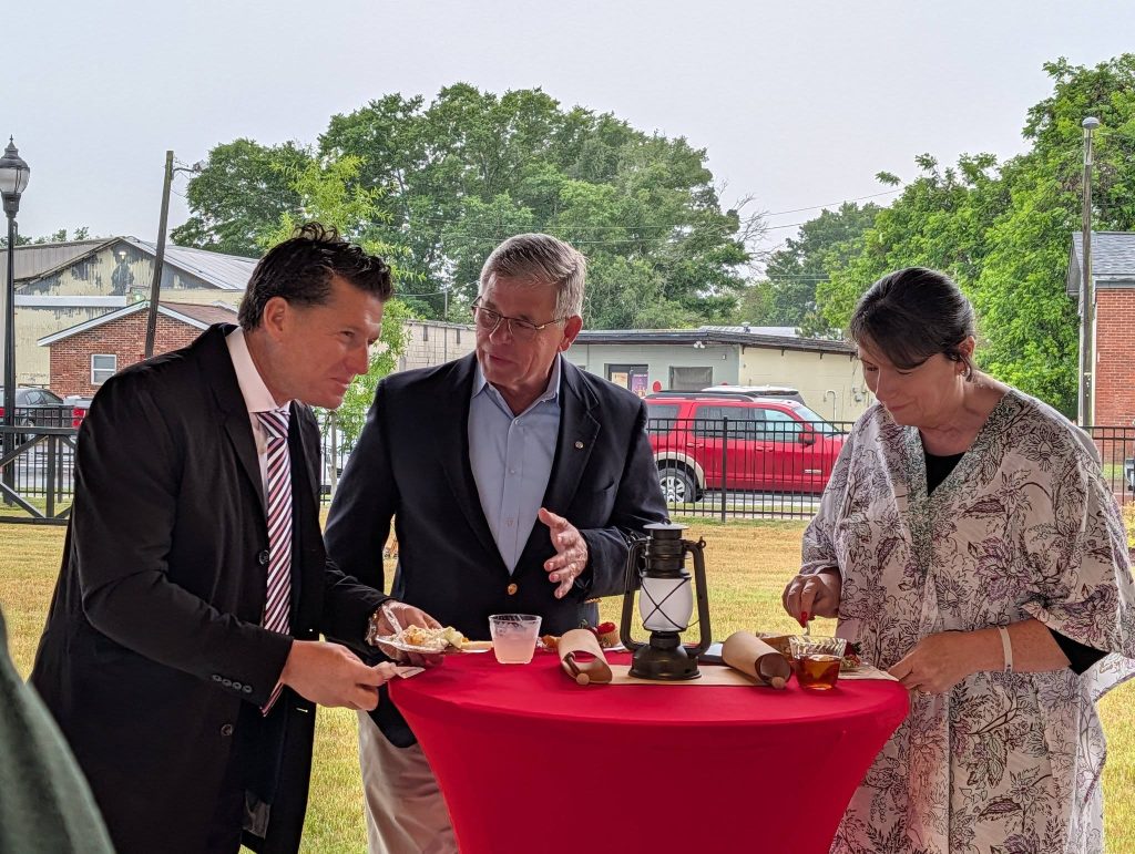 Three adults stand around a red table outdoors, eating and talking at a casual event, with buildings and trees in the background.
