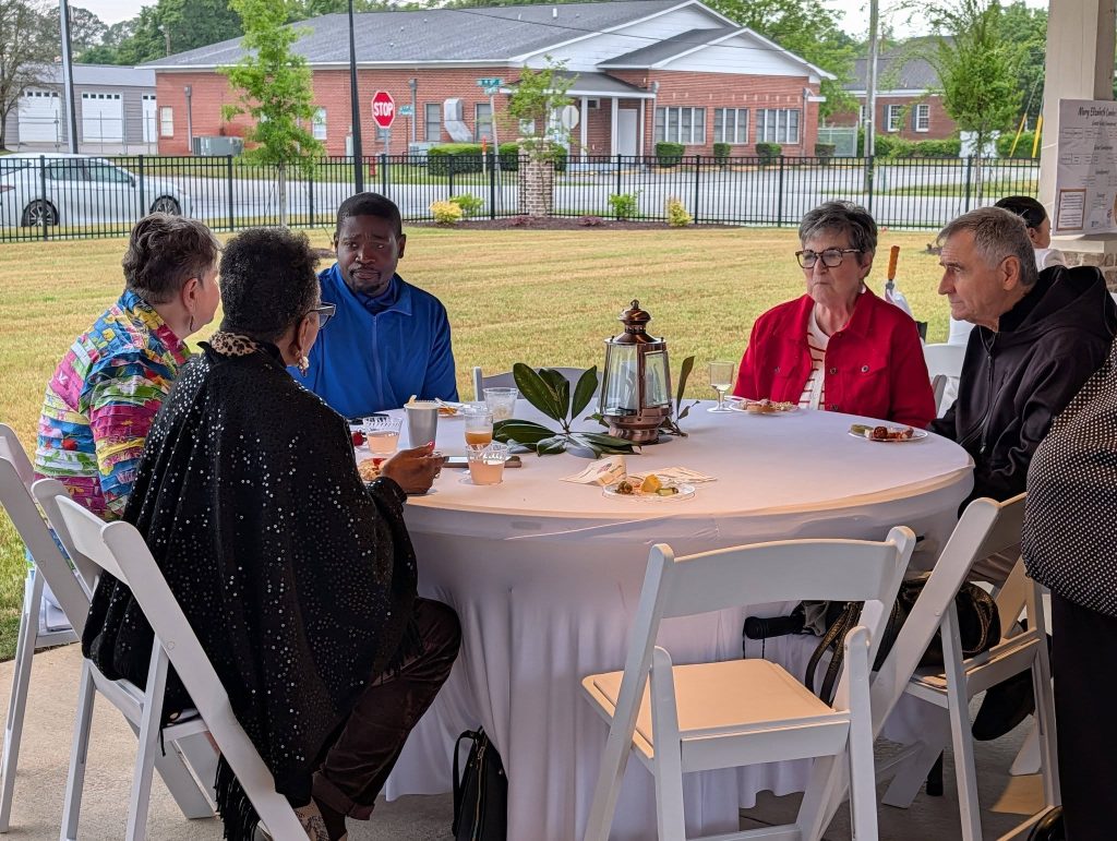 Six people sit around a round table outdoors, engaged in conversation, with food and drinks on a white tablecloth, and buildings visible in the background.