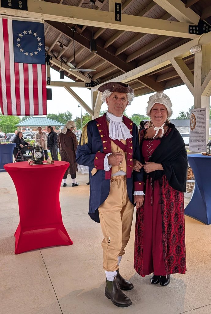 Two adults in colonial-era costumes stand under a pavilion with a historical American flag and people in period attire in the background.