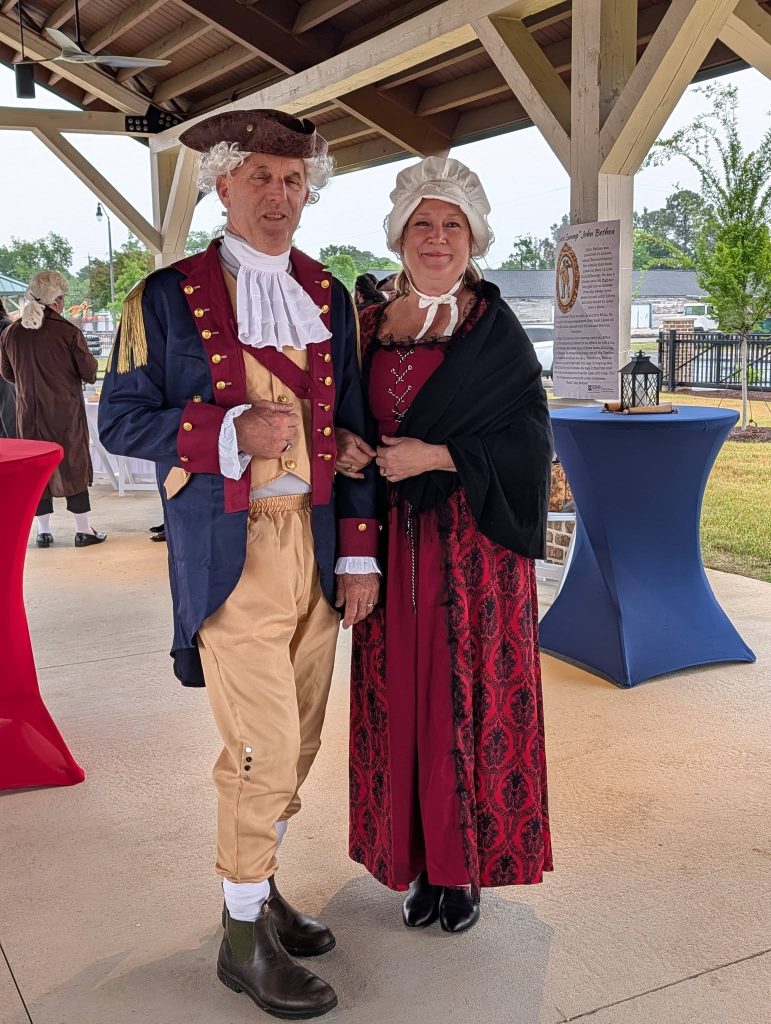 A man and woman dressed in colonial-era costumes stand under a pavilion, smiling for the camera at a historical reenactment event.