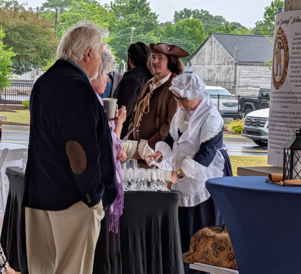 People dressed in historical costumes stand around a table with glasses, engaging in conversation at an outdoor event with informational displays in the background.