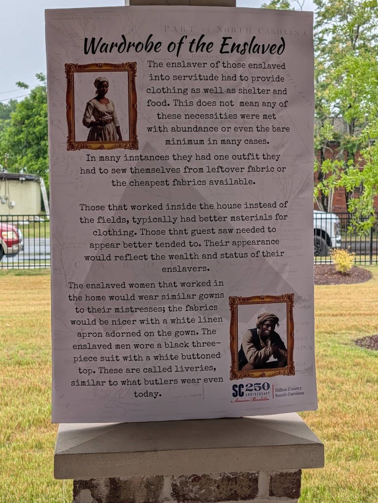 Exhibit sign titled "Wardrobe of the Enslaved," describes clothing conditions for enslaved people, with images of a woman and a man in period dress, outdoors near grass and trees.
