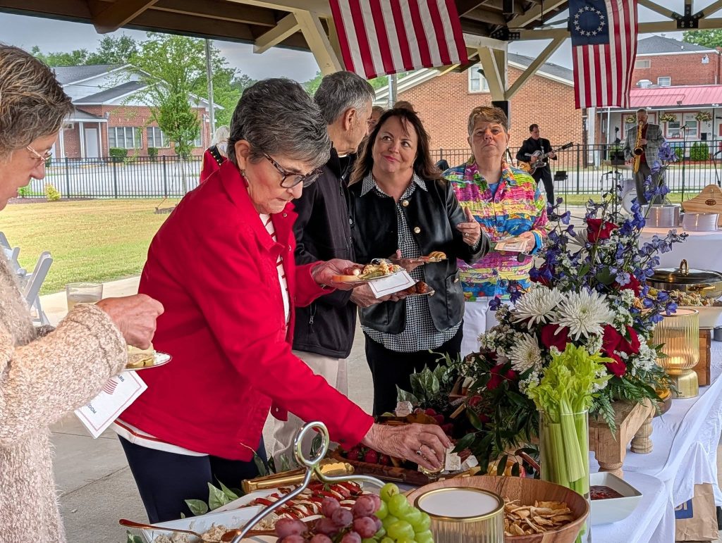 People serve themselves food at a buffet table adorned with flowers under a pavilion decorated with American flags.