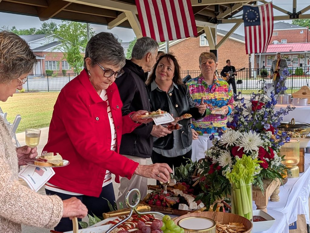 People serve themselves food from a buffet table with flowers, under American flags at a social gathering, while musicians play in the background.