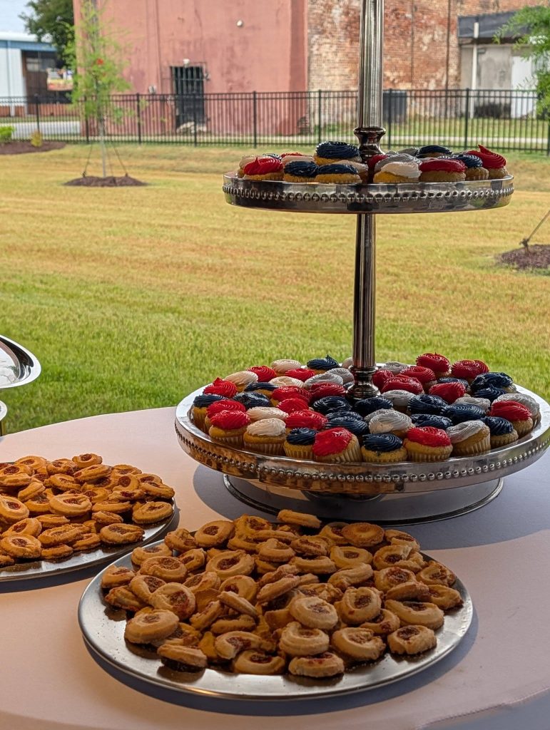 Plates of cookies and a tiered tray with mini cupcakes decorated with red, white, and blue frosting are set on a table outdoors.