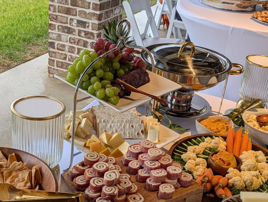 A table with assorted appetizers including grapes, cheeses, crackers, vegetables, dips, and rolled meat snacks, set for an outdoor event with white tablecloths.