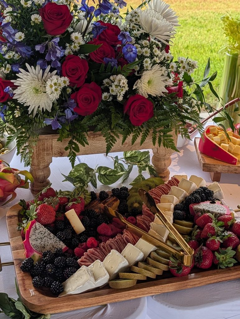 Charcuterie board with cheeses, berries, dragon fruit, kiwi, and salami, set in front of a vibrant flower arrangement with red roses, white daisies, and purple flowers.