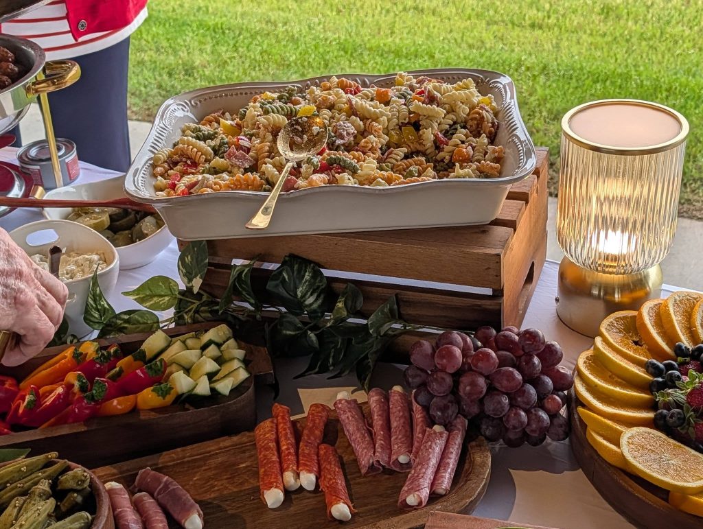 A buffet table with pasta salad, fresh fruits, cured meats, vegetables, dips, and a decorative lamp, set outdoors with greenery in the background.