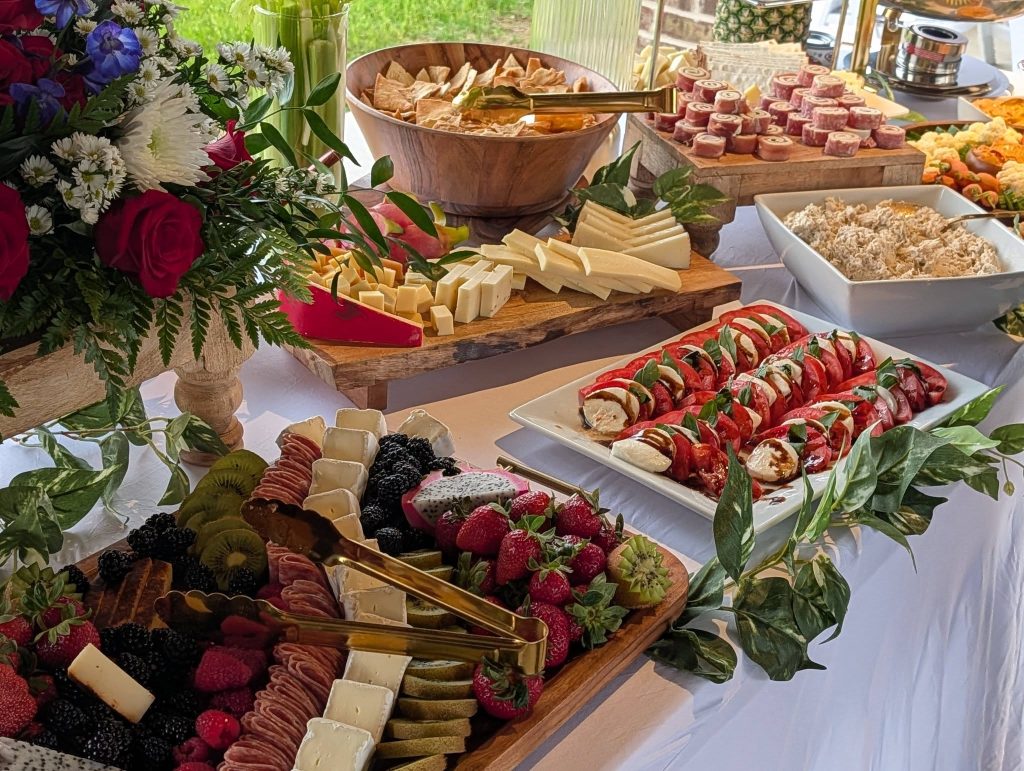 A buffet table with assorted cheeses, meats, fresh fruit, Caprese salad, dip, crackers, and flowers, arranged for a festive gathering.
