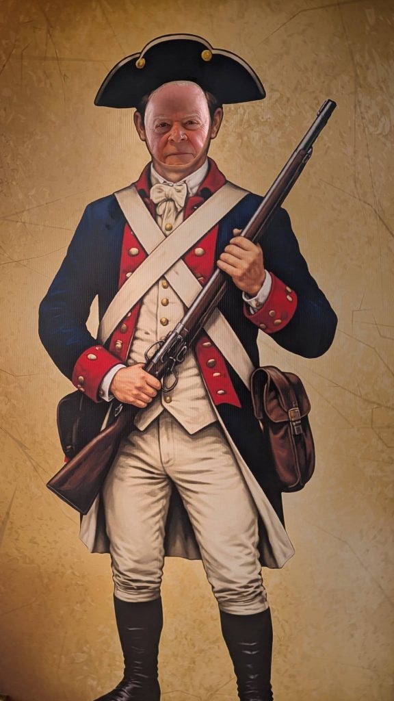 A man's face in a cutout board of a Revolutionary War soldier, wearing a uniform and holding a musket.