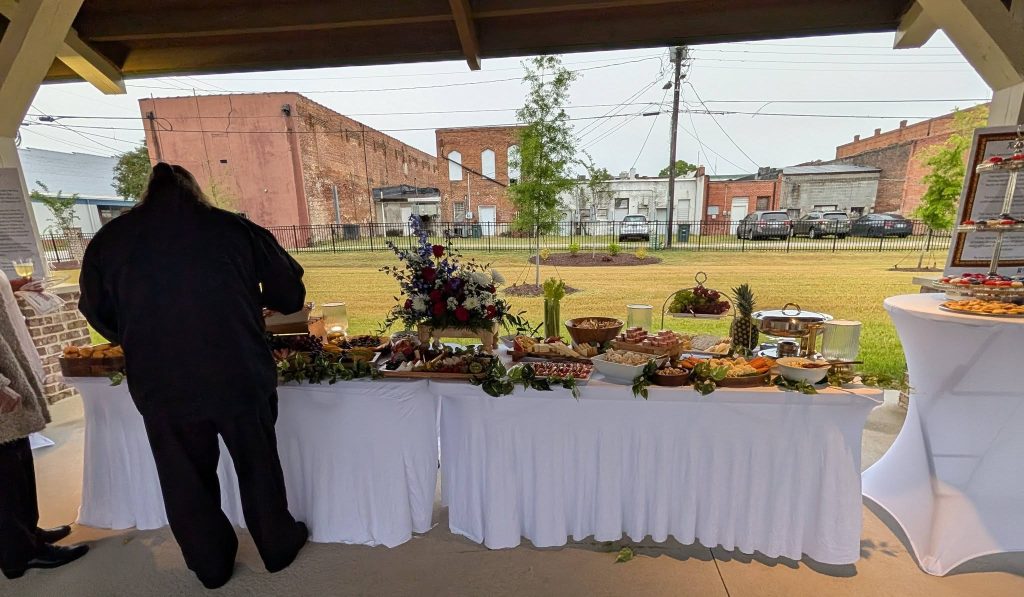 Buffet table with various appetizers and snacks, decorated with a floral arrangement, outdoors under a pavilion with people serving themselves. Brick buildings and parked cars in the background.