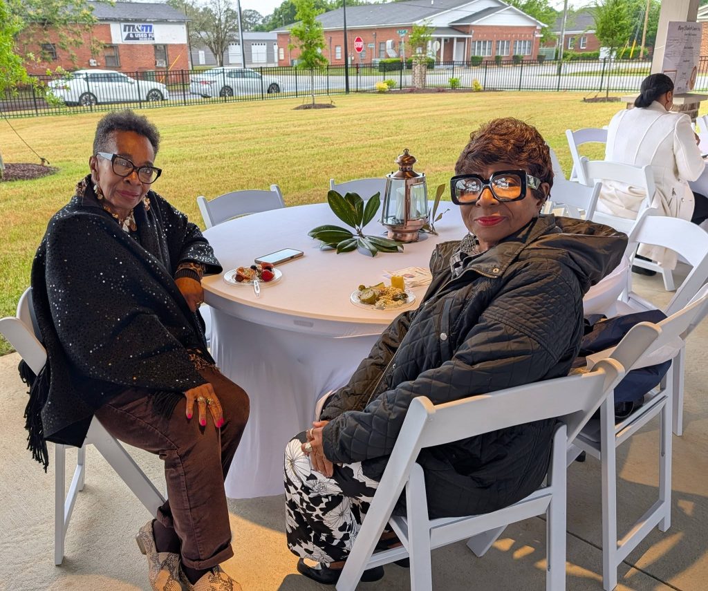 Two women with glasses sit at an outdoor table with white chairs, enjoying food and conversation, with greenery and buildings visible in the background.