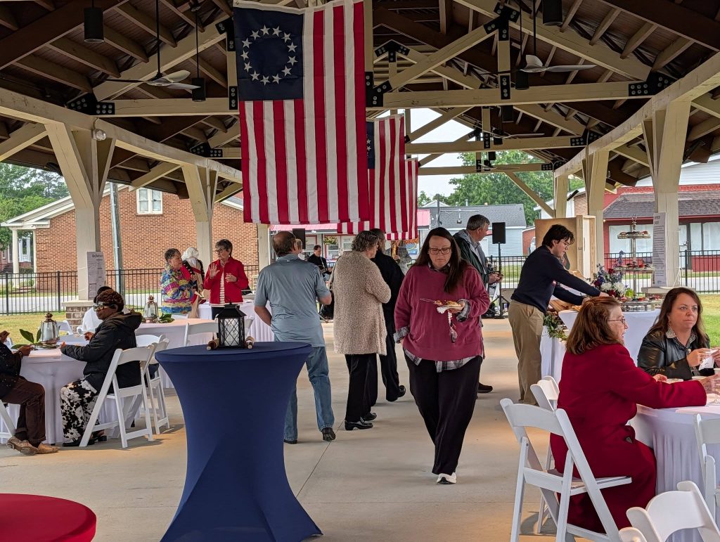 People gather under a pavilion decorated with American flags, sitting and standing around tables, enjoying food and conversation during an outdoor event.