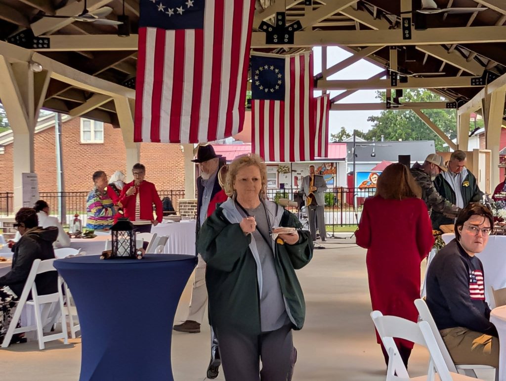 People attending an indoor event with American flags hanging overhead, various attendees are talking, eating, and sitting at tables under a pavilion.