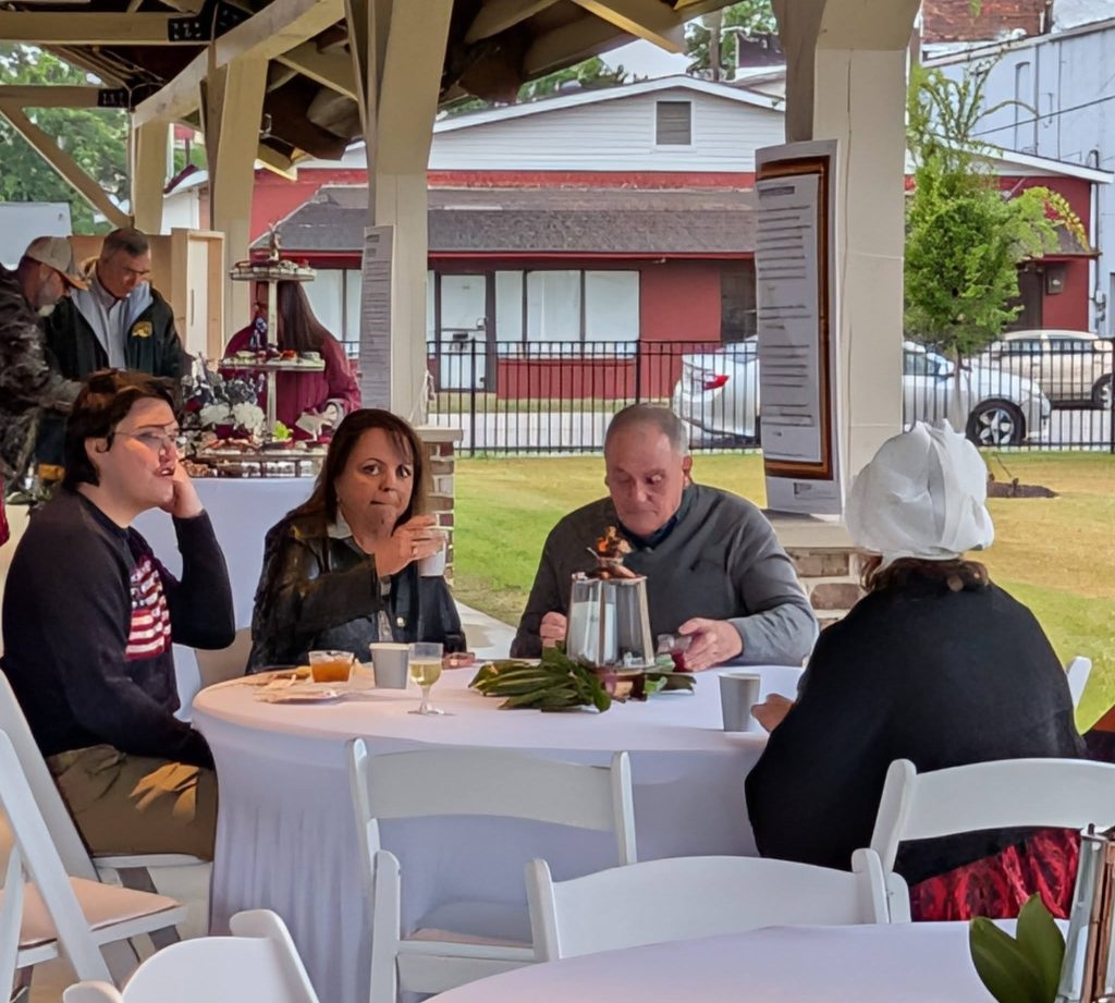 Four people sit at a round table under a pavilion, drinking and eating at an outdoor event with white tablecloths and decorations.