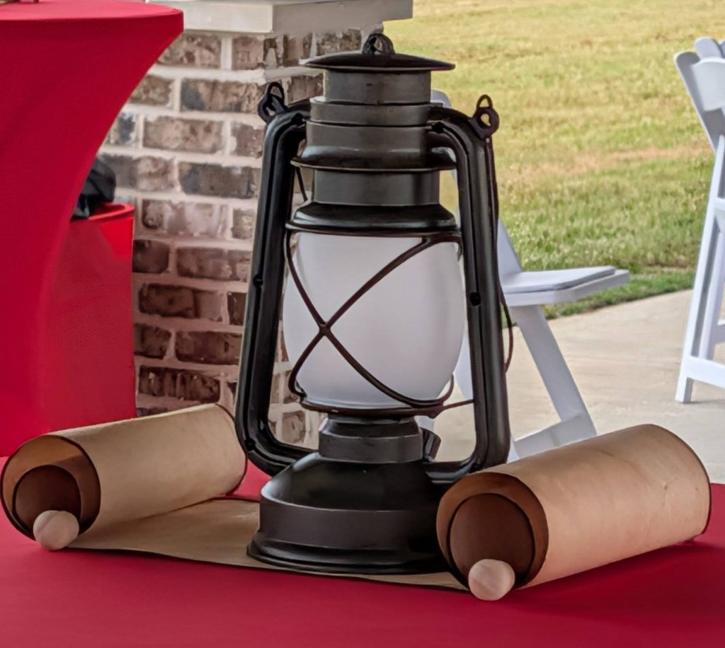 A vintage lantern sits on a red table, flanked by two scrolls. Brick wall and outdoor grass are visible in the background.