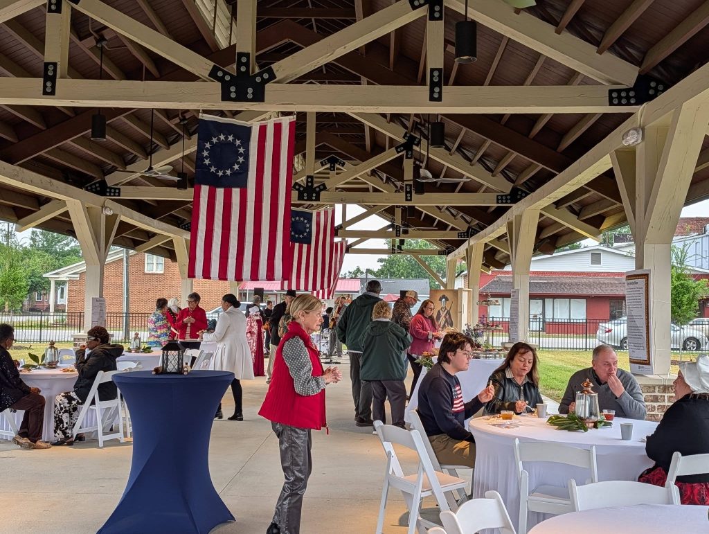 People gathered under a pavilion decorated with colonial American flags, sitting and standing around tables, enjoying food and conversation at a community event.