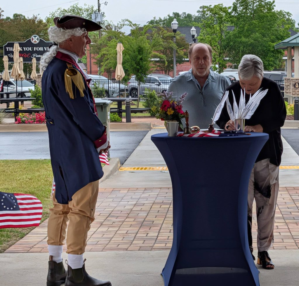 Three adults participate in a patriotic-themed outdoor ceremony, one in historical attire, while a woman signs documents on a table decorated with quill pens and flowers.