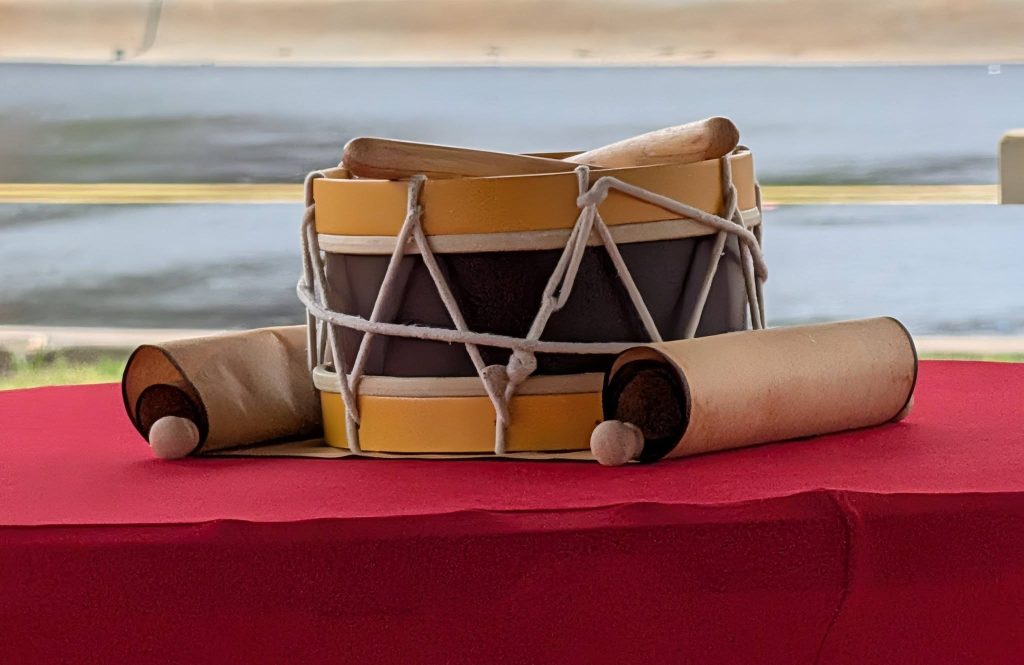A small drum with drumsticks and two rolled scrolls on a red cloth-covered table near a body of water.
