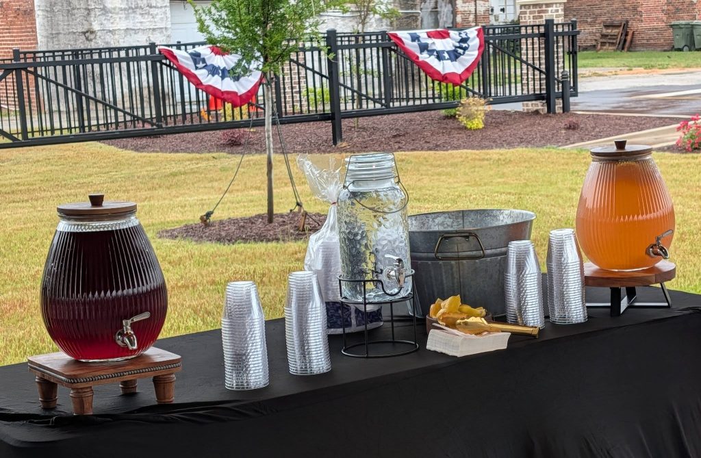 Outdoor refreshment table with large drink dispensers, plastic cups, ice container, and patriotic decorations in the background.