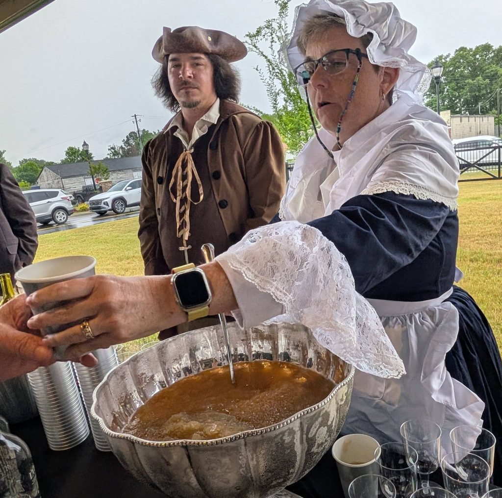 Two people in colonial-style costumes serve punch from a large bowl outdoors, with cars and grass visible in the background.