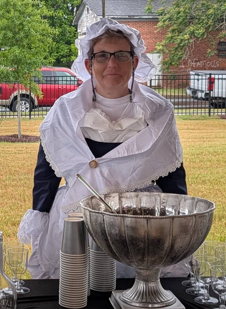 Person in colonial-style attire stands behind a table with a large silver punch bowl, plastic cups, and glasses outdoors.