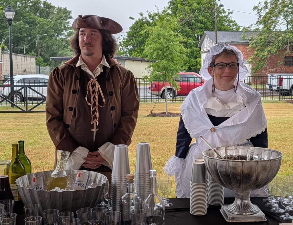 Two people in colonial costumes stand behind a refreshment table outdoors, featuring bottles, cups, glassware, and a large silver punch bowl.