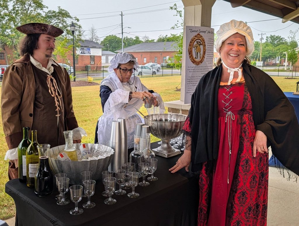 Three people in colonial-style costumes serve drinks at a table with bottles, glasses, and ice, during an outdoor historical reenactment event.