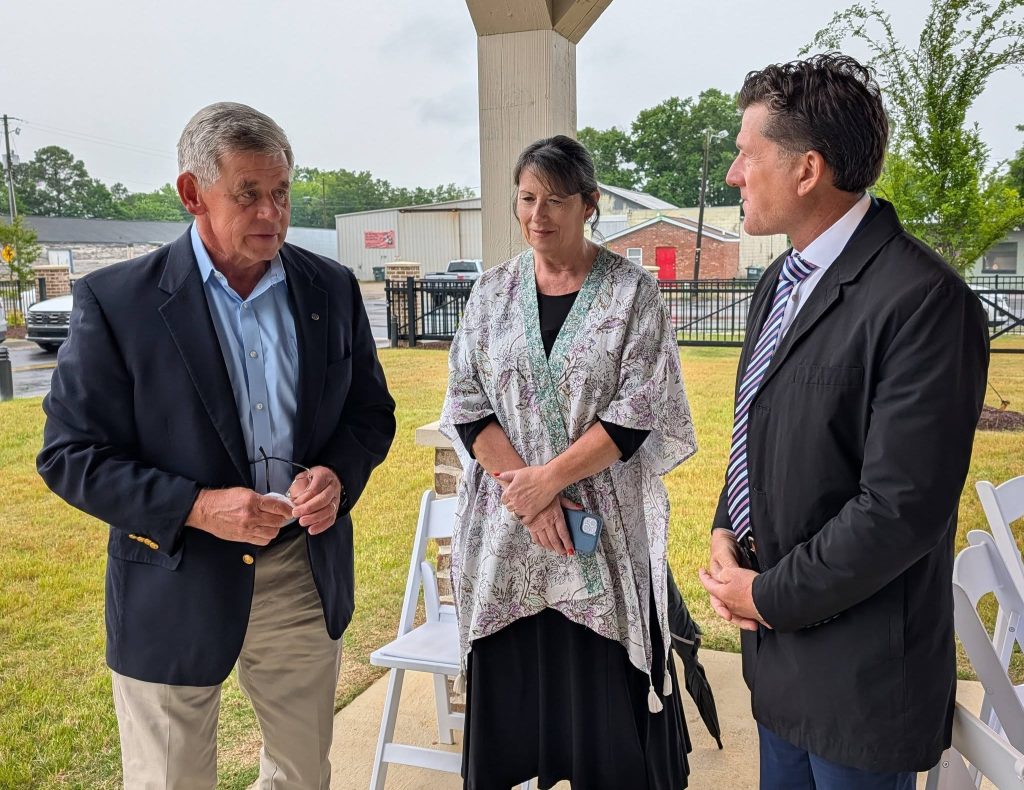 Three people in business attire are standing outside under a covered area, having a conversation near white folding chairs on a cloudy day.
