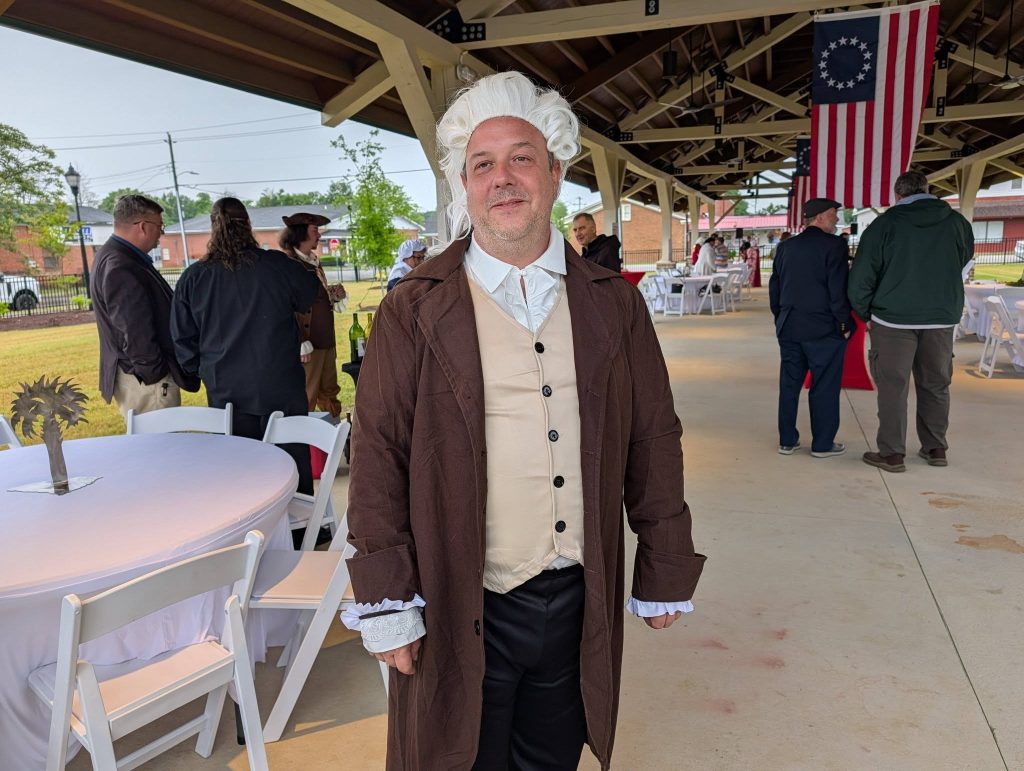 A man in historical colonial attire stands under a pavilion decorated with an early American flag, surrounded by people at an event with round tables and chairs.