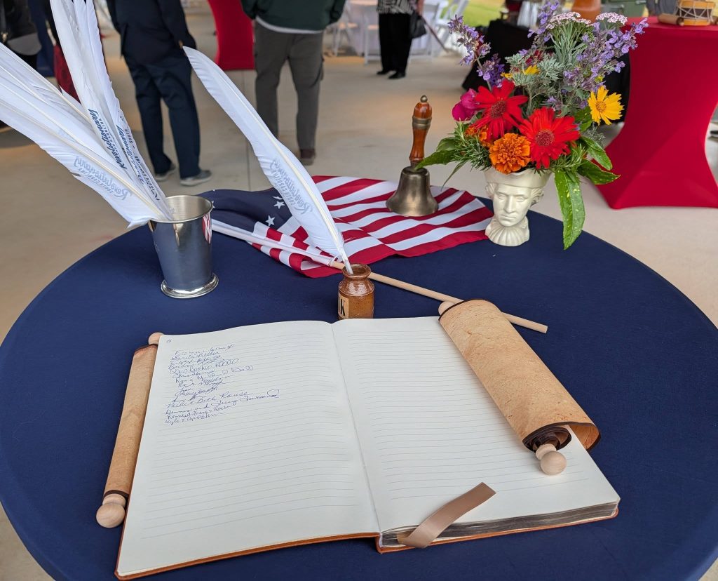 A table with a guest book, feather quill pens, American flag, bell, flower arrangement, and scroll, with people and red-covered tables in the background.