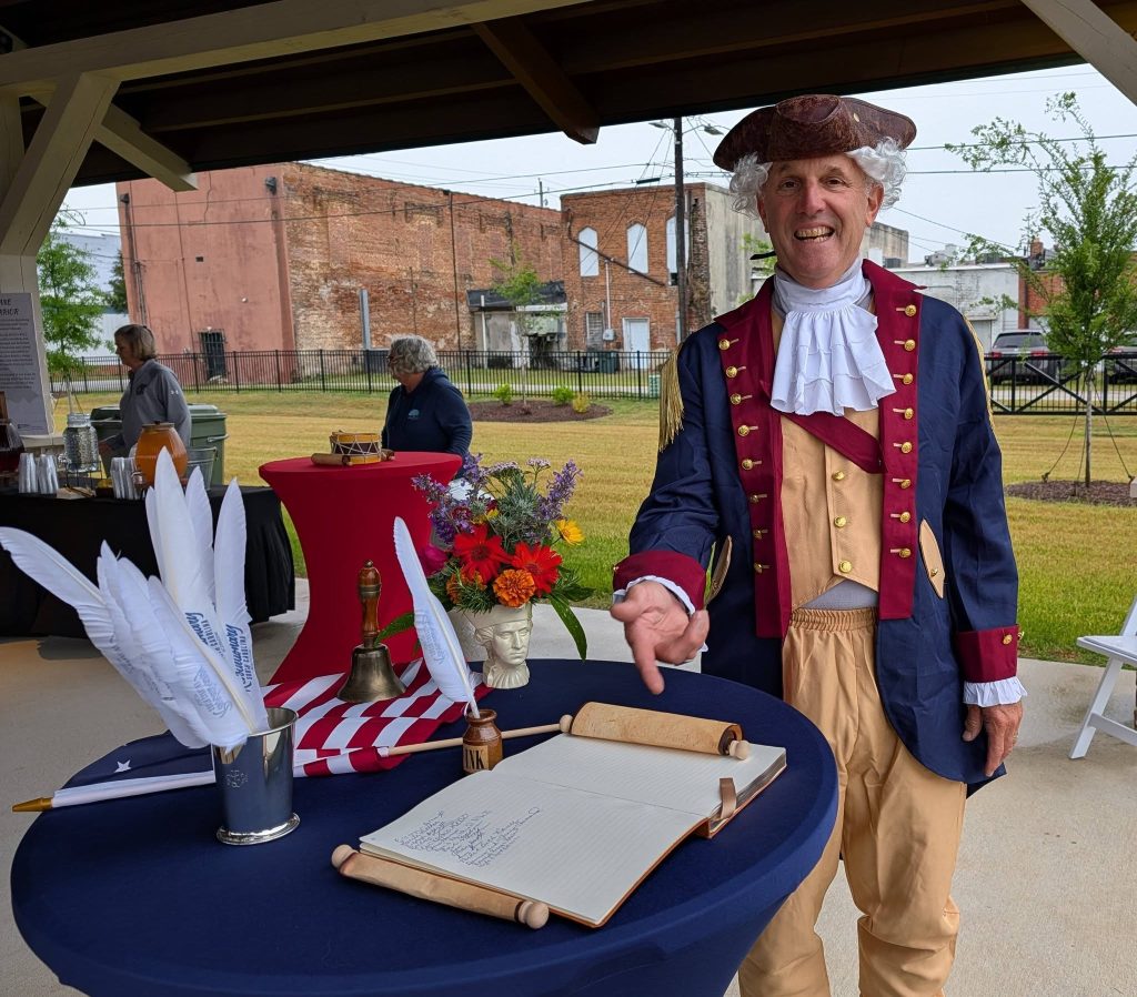 A man in historical costume stands by a table with a large open book, feather quills, flowers, and patriotic decorations at an outdoor event.