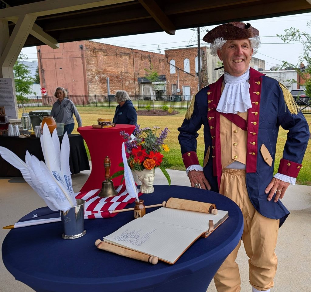 Man in Revolutionary War costume stands by decorated table with quill pens, open guest book, bell, and flowers at an outdoor event.