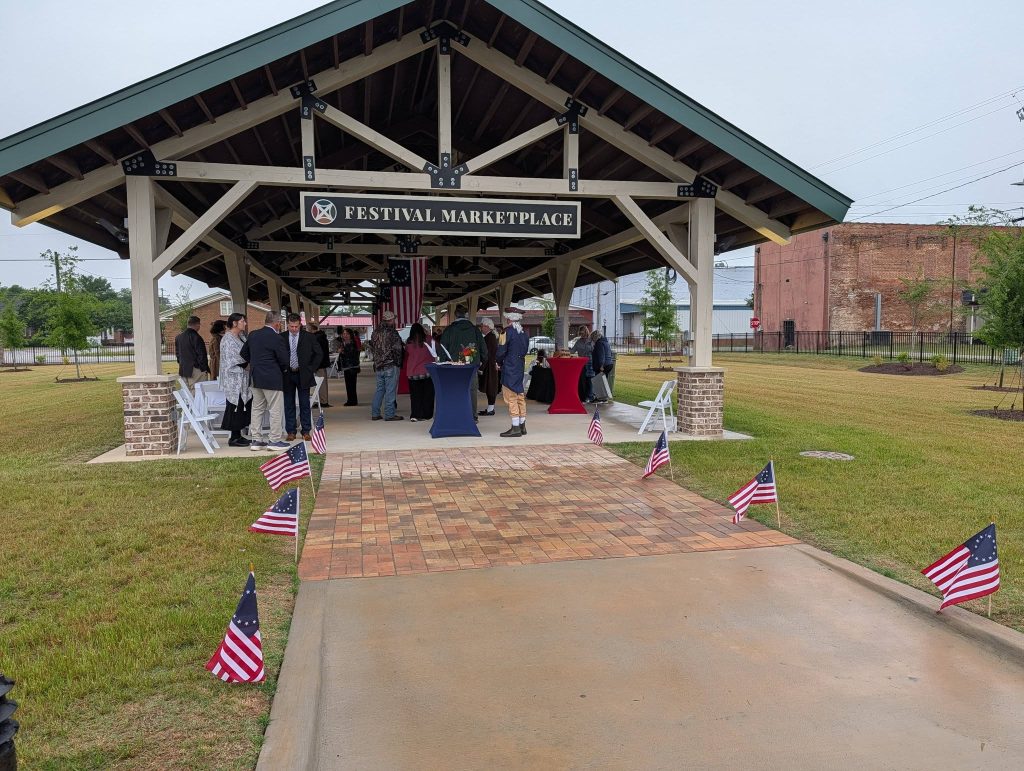 People gather under a pavilion labeled "Festival Marketplace," surrounded by American flags and chairs on a cloudy day, with grass and brick buildings in the background.