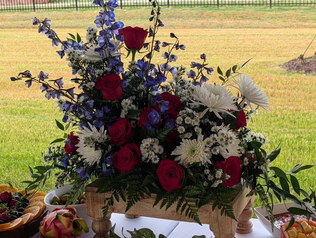 A wooden box centerpiece filled with red roses, white chrysanthemums, purple flowers, and greenery, displayed on a table with fruit dishes outdoors.