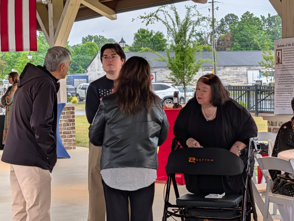 Four people converse under a pavilion, one woman using a walker, with an American flag above and trees and parked cars visible outside.