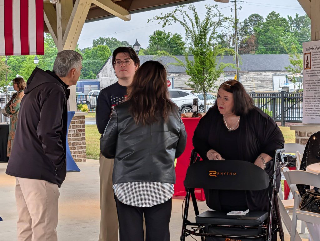 Four people converse at an outdoor covered event, with one woman using a walker. American flag and another attendee are visible in the background.