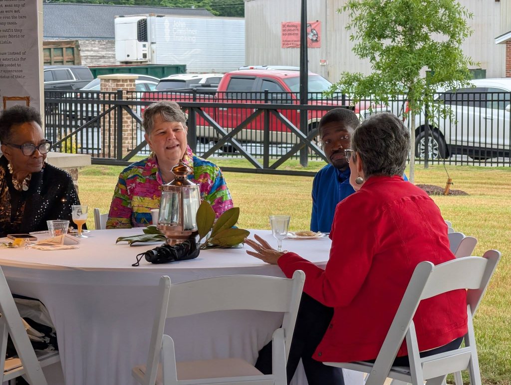 Four adults sit around a table outdoors, engaged in conversation, with drinks and a lantern on the table. Cars and greenery are visible in the background.