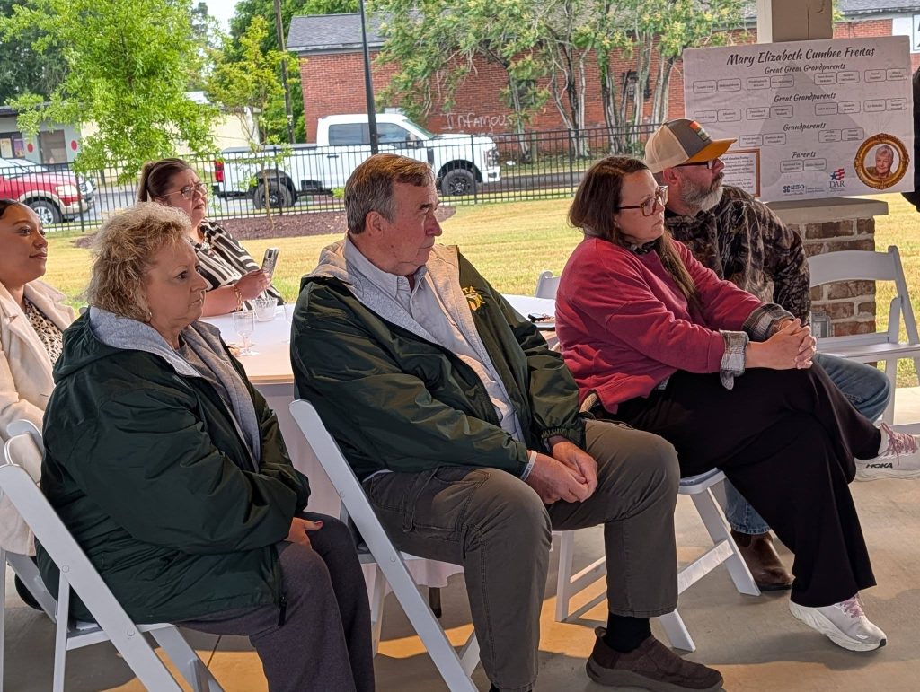 A group of people sit attentively on white chairs at an outdoor covered event, with a family tree poster visible in the background.