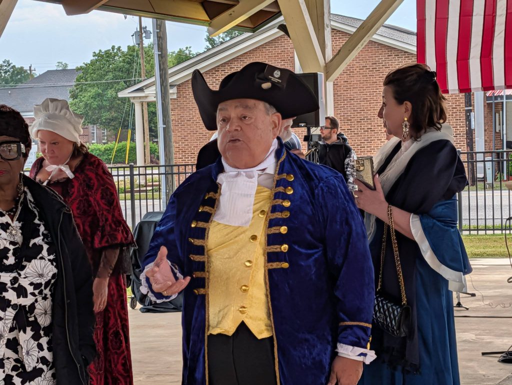 A group of people wearing historical costumes converse under a pavilion, including a man in a tricorn hat and blue coat. An American flag hangs in the background.