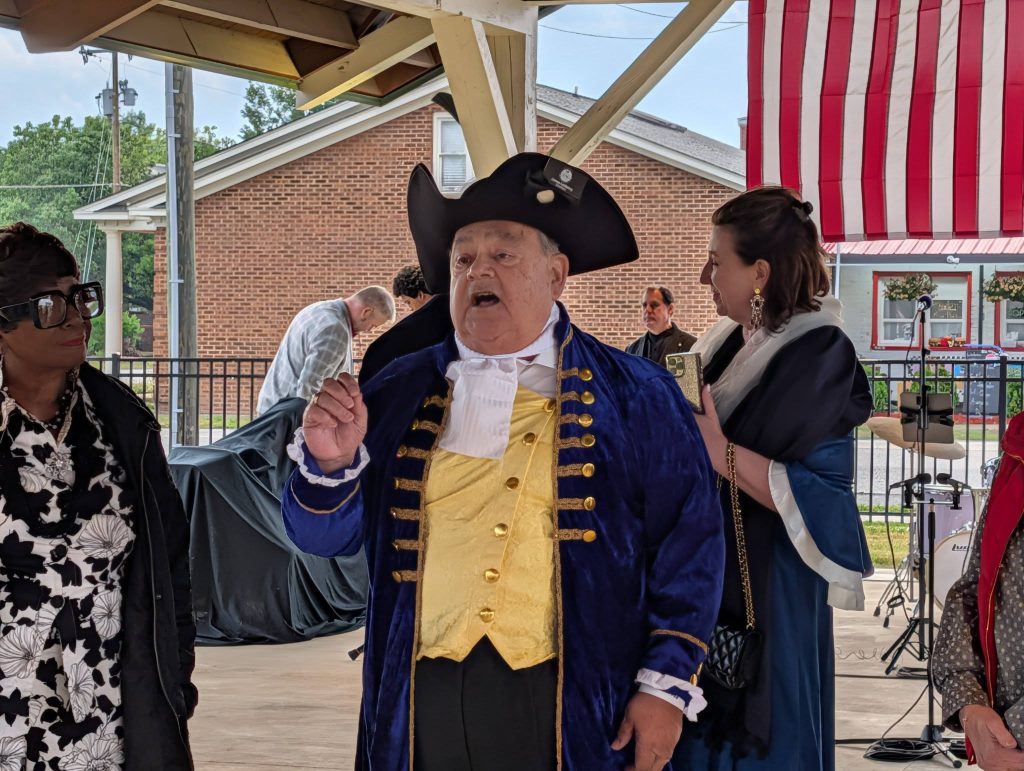 Man in historical colonial costume speaks at an outdoor event, with other people around him and a large American flag in the background.