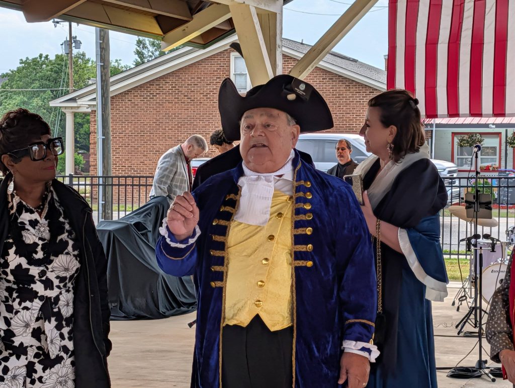 A man in colonial costume speaks at an outdoor event, surrounded by people in modern and period clothing, with an American flag hanging in the background.
