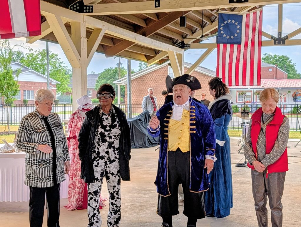A group of people, some in historical costumes, stand under a pavilion with a colonial American flag hanging in the background.