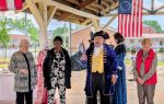 Four adults stand under a pavilion, one dressed in colonial attire, with a historical American flag hanging in the background.