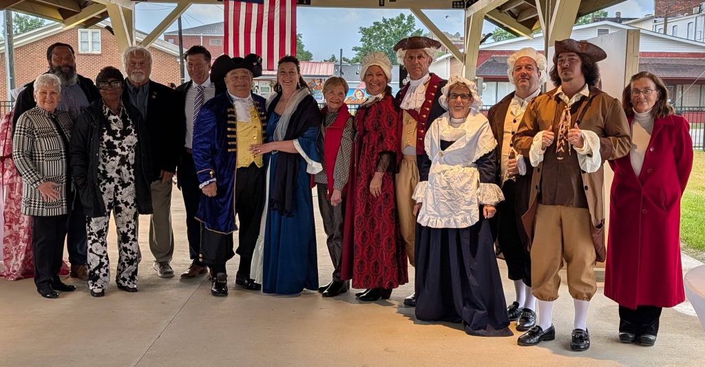 A group of people pose together, some dressed in modern clothes, others in historical colonial costumes, under a pavilion with an American flag in the background.