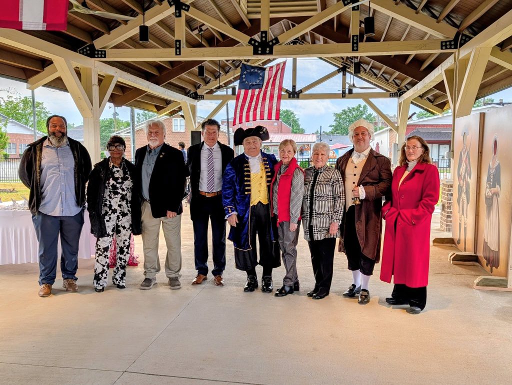 A group of people pose under a pavilion, some in colonial costumes, with an American flag hanging above them.