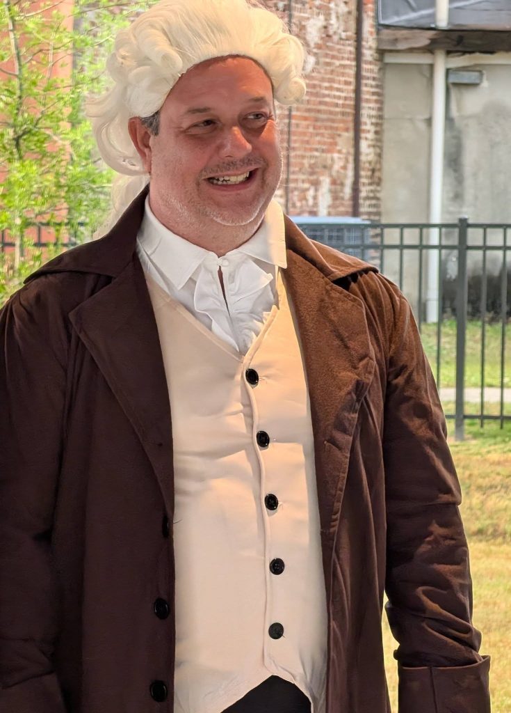 Man wearing a white powdered wig, white shirt, cream waistcoat, and brown coat, dressed in historical or colonial-style costume outdoors.