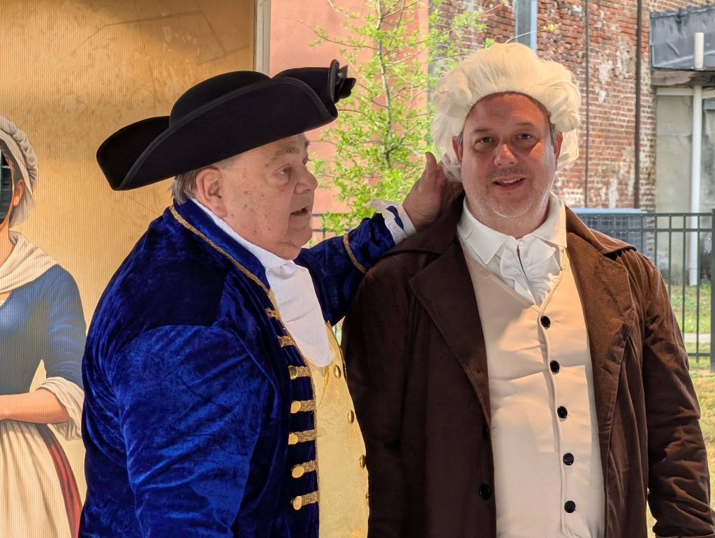 Two men dressed in historical colonial costumes, one with a tricorn hat and velvet coat, the other with a white wig and brown coat, posing indoors.