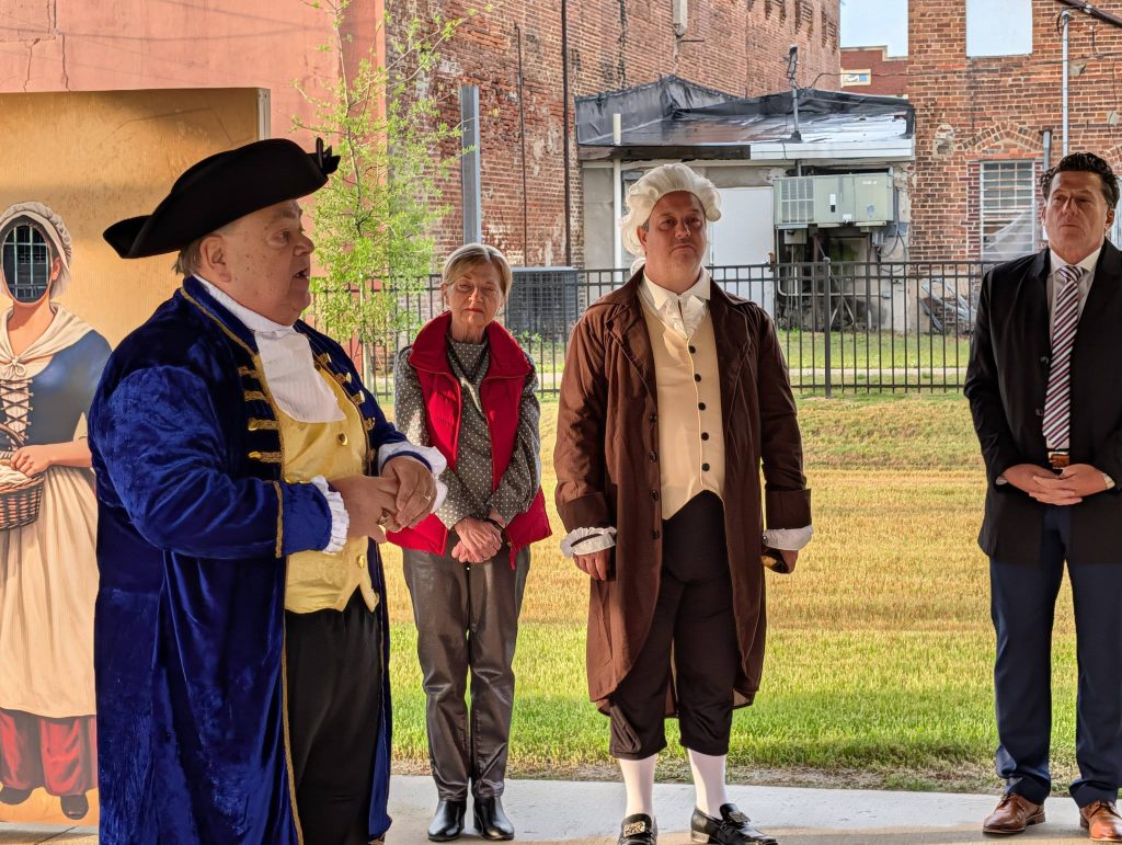 Four adults stand outdoors, two in historical colonial costumes, one in a modern suit, and one in casual clothing, with a cutout in the background.