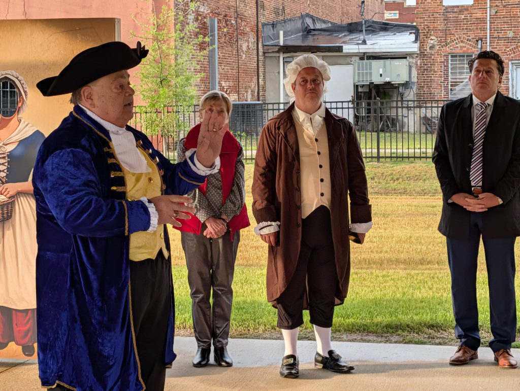 Four adults stand outdoors, two dressed in historical colonial costumes, and two in modern clothing, with a brick building and signage in the background.